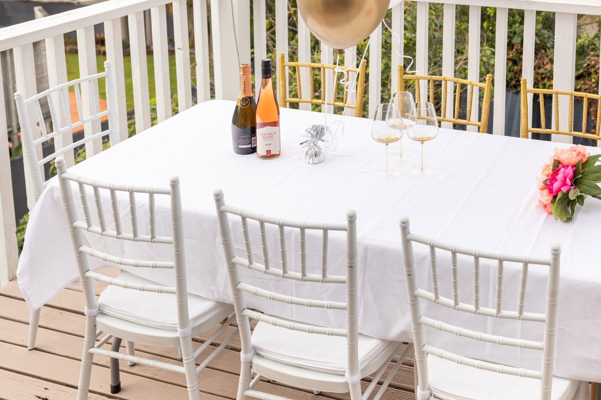 Long white-clothed table on a timber deck laid with champagne, glassware and pink flowers, surrounded by white Chiavari chairs