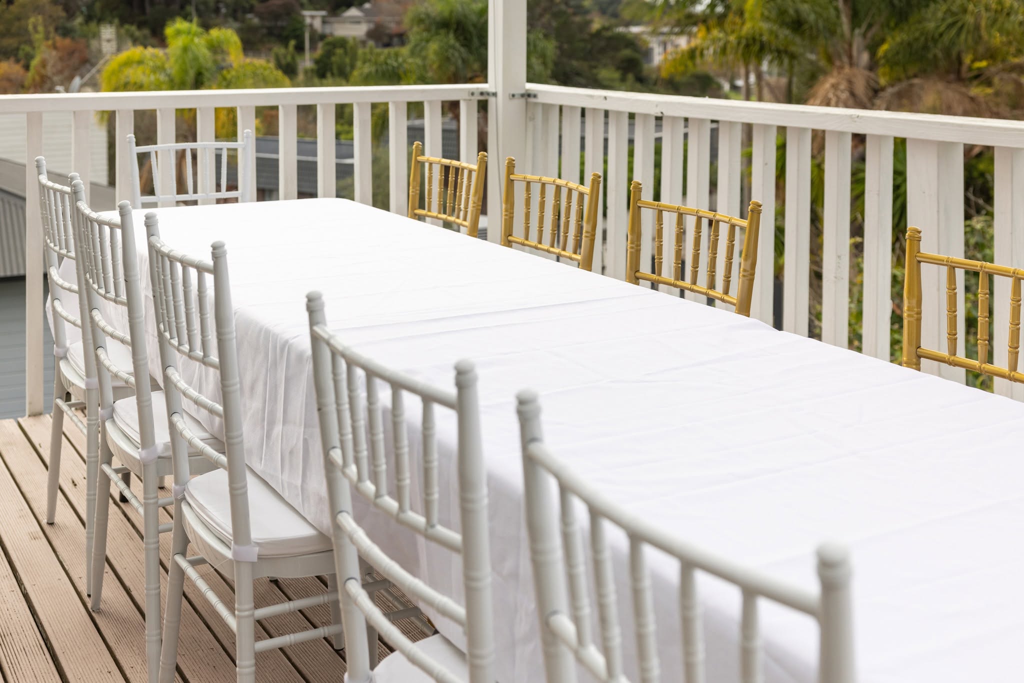 Two trestle tables joined end to end under white linen on a deck, with white Chiavari chairs tucked along one side