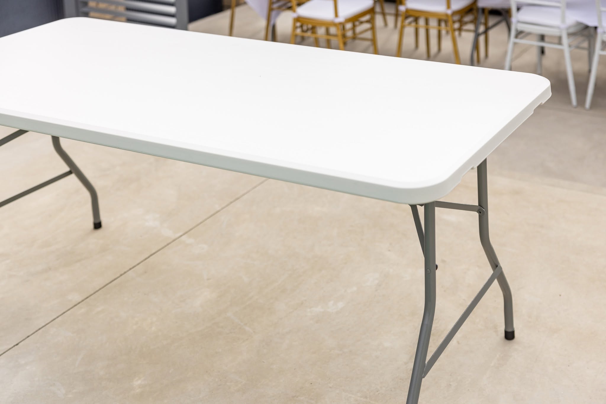 White folding trestle table set up on a concrete patio with its grey steel legs open, chairs stacked behind